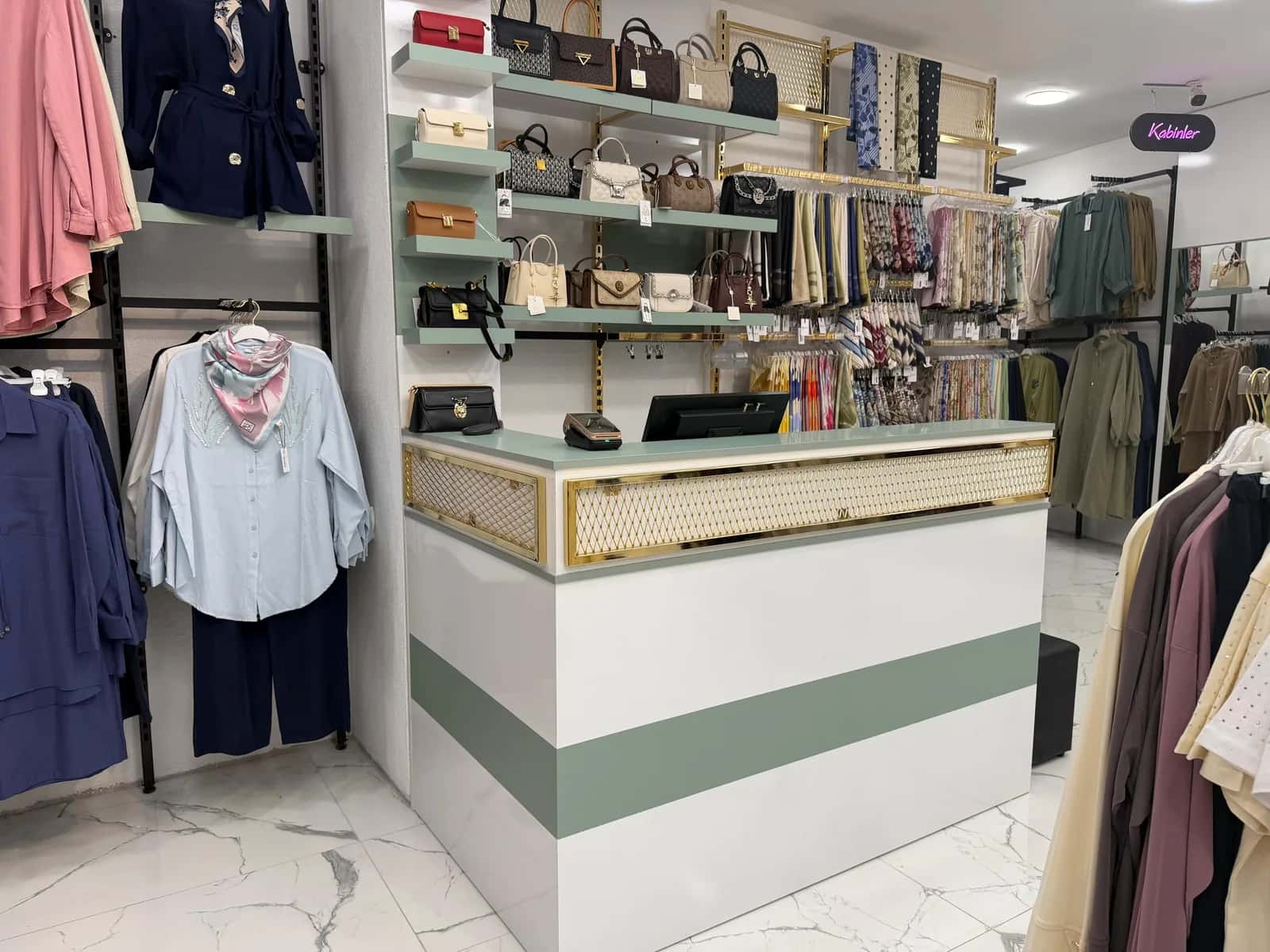 Armira mağazasında beyaz gövdeli, yeşil bantlı ve altın kafes panelli kasa bankosu, arkada çanta rafları ve eşarp duvarı