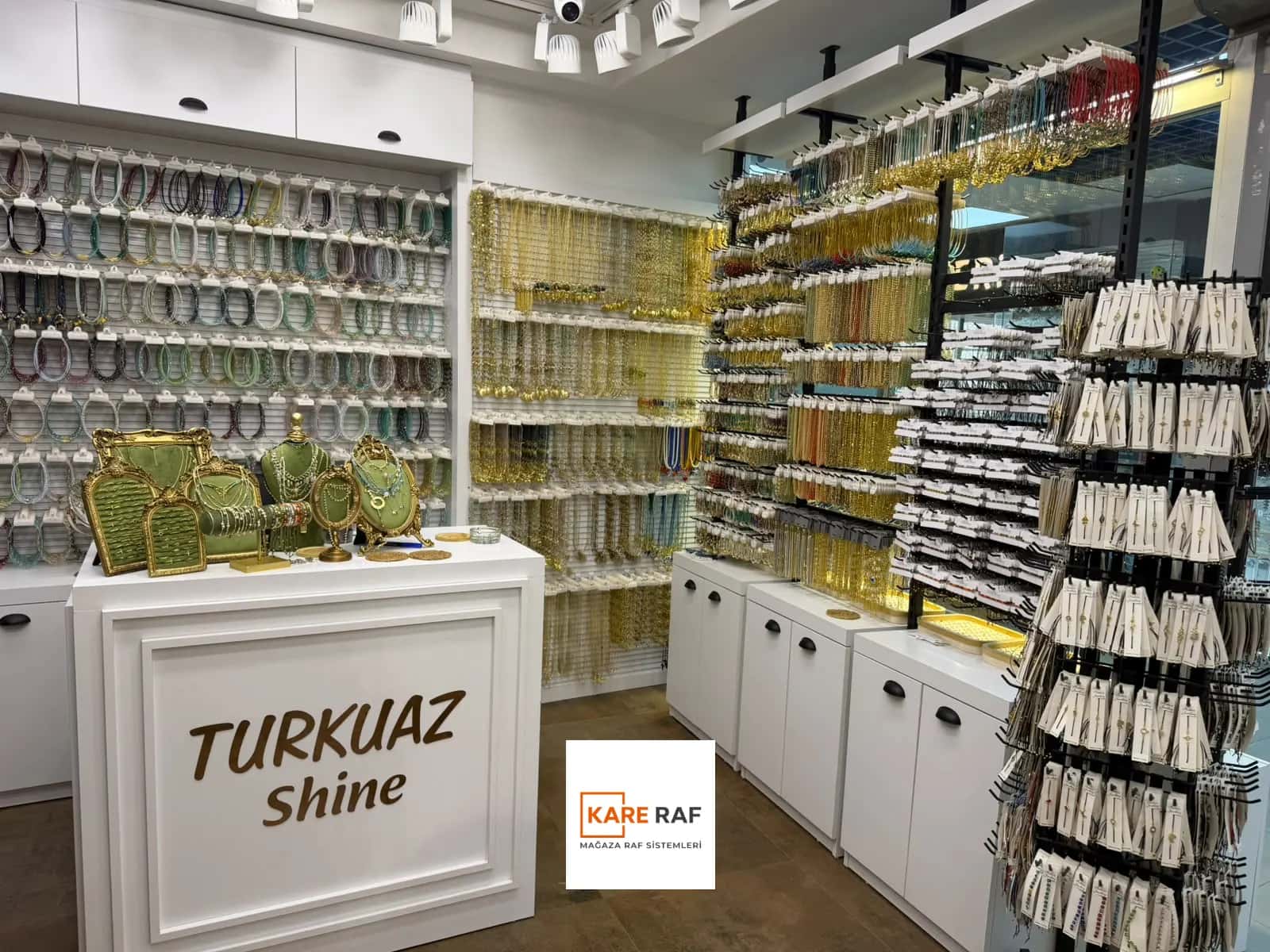 Turkuaz Shine yazılı beyaz kasa bankosu; arkada kanallı panoda bileklikler, yanda kolye raflı duvar