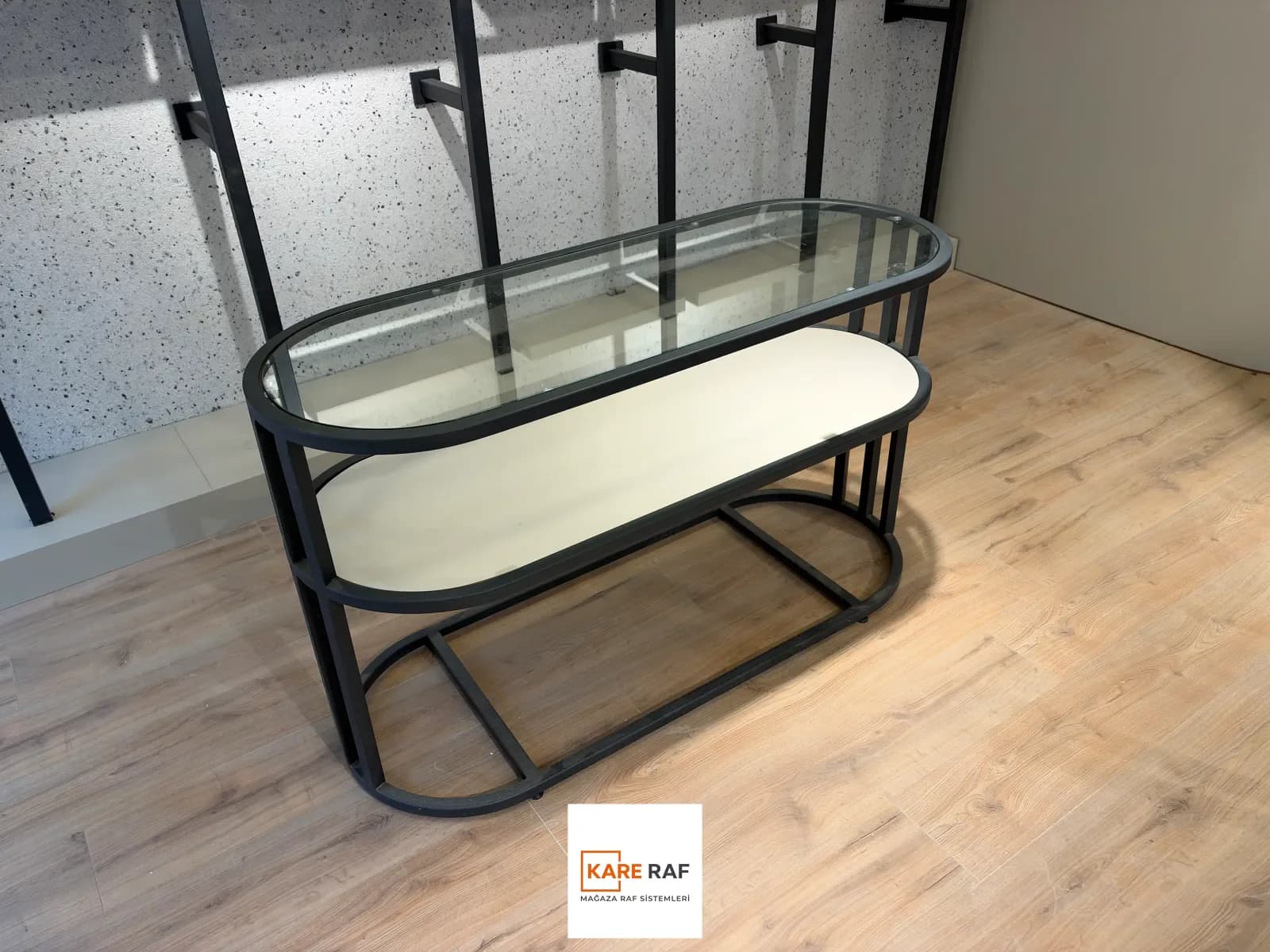 Oval cam üstlü, antrasit metal çerçeveli orta camlı teşhir bankosu; altında kum beji tabla