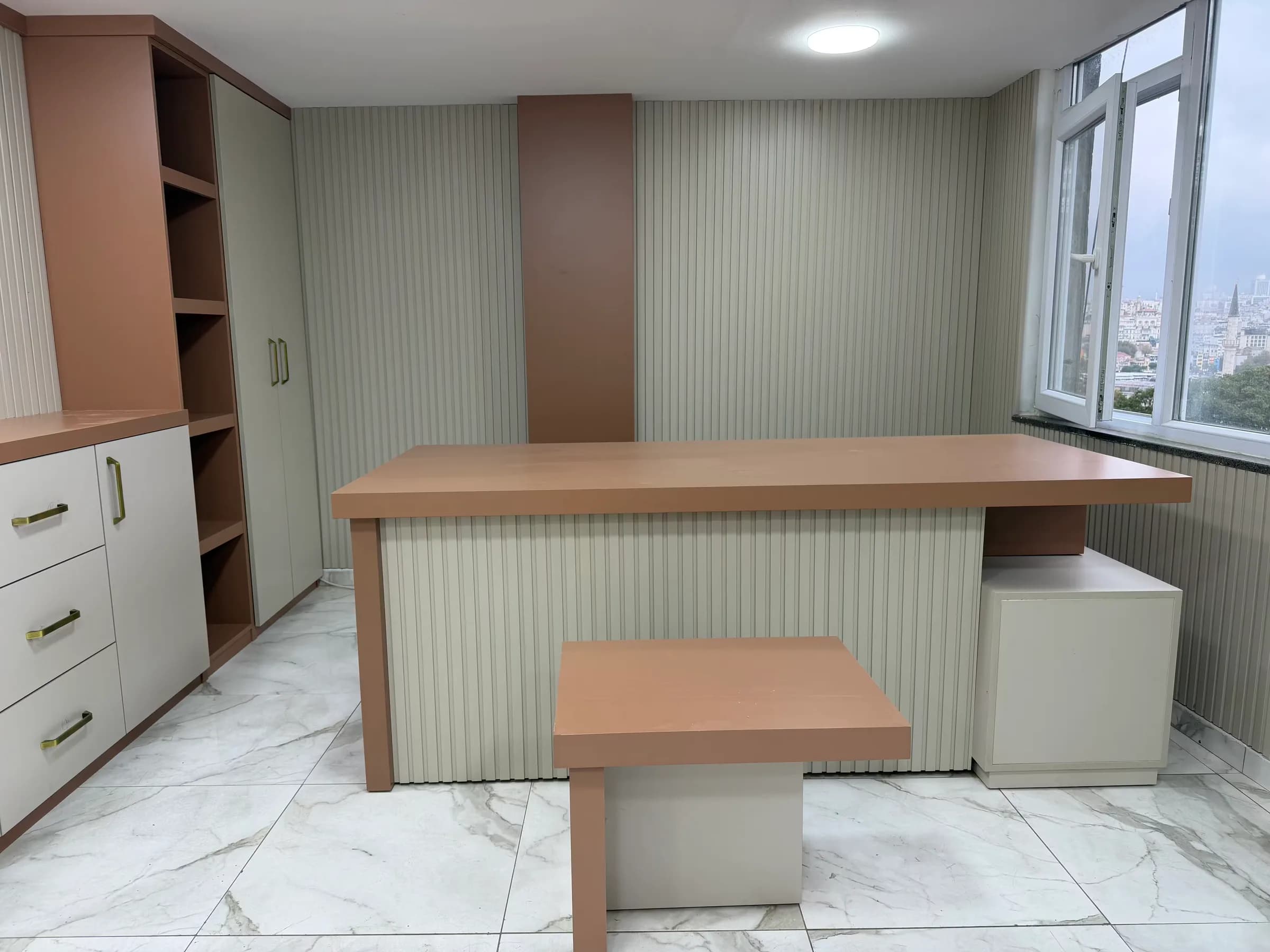 Önü krem ahşap lambri kaplı, kiremit tablalı çalışma masası, sağında krem dolap, önünde küçük masa