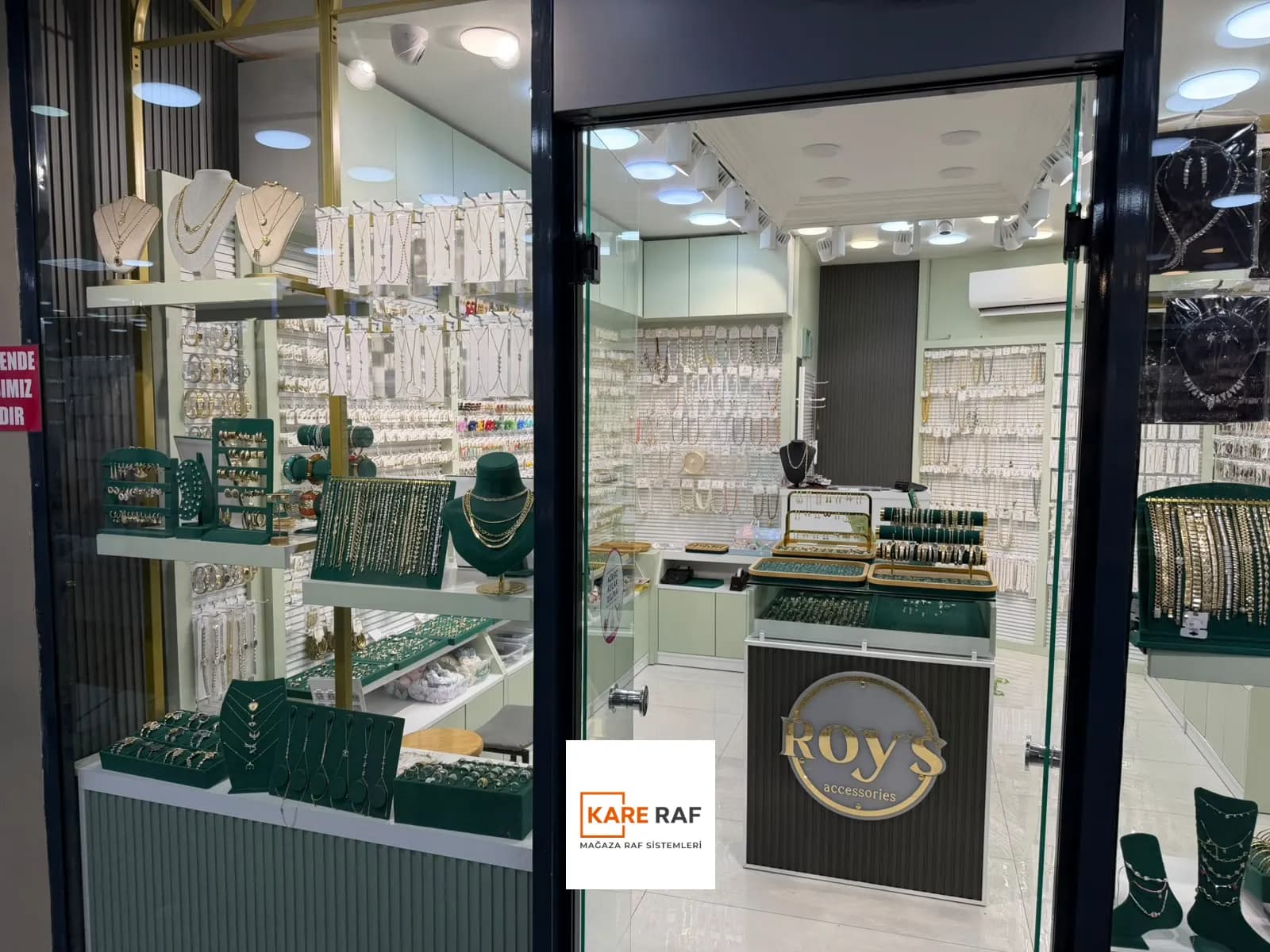 Sol vitrin ve giriş: raflarda kolye büstleri, içeride kanallı pano duvarlar ve logolu orta stand