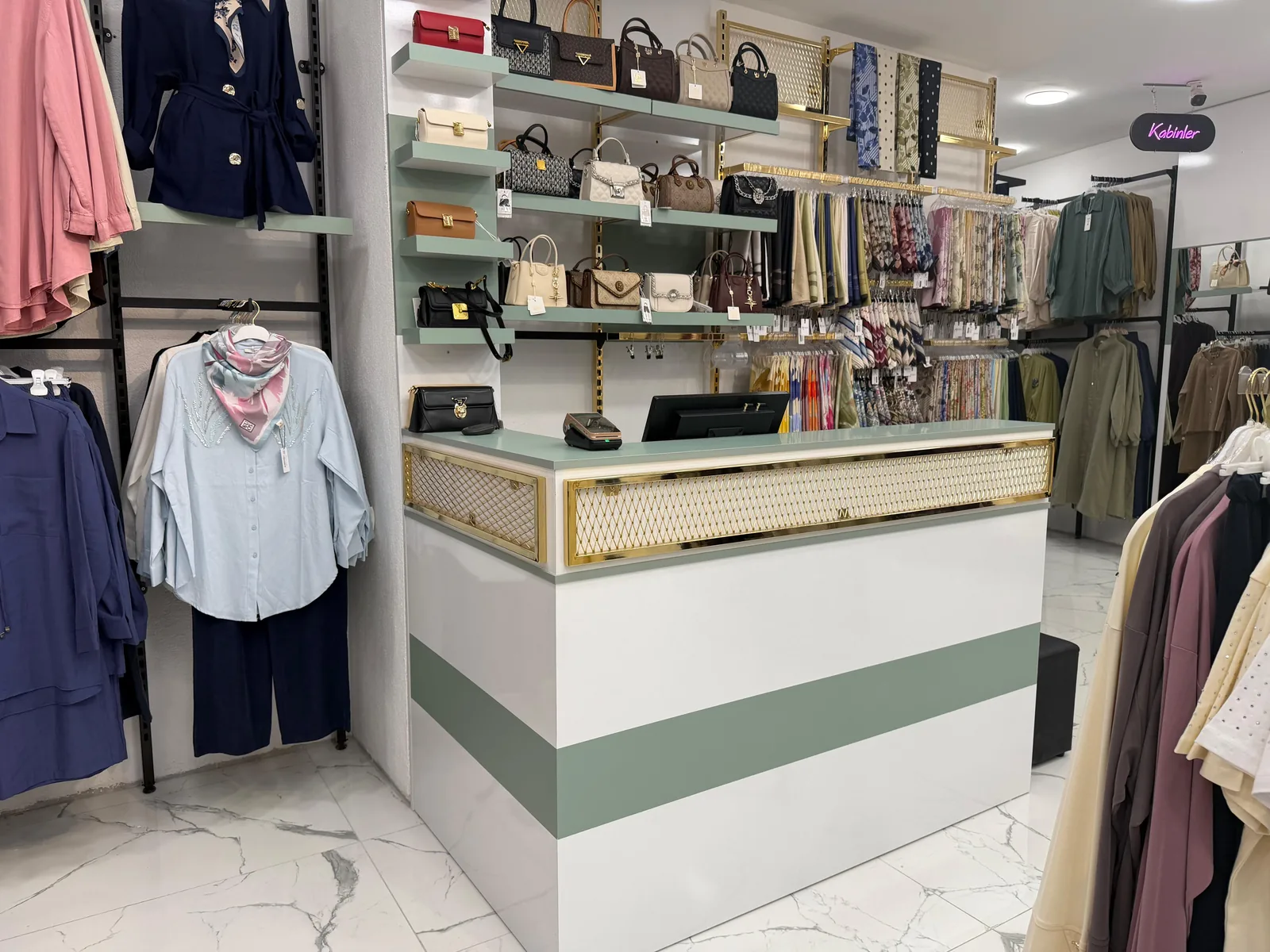Armira mağazasında beyaz gövdeli, yeşil bantlı ve altın kafes panelli kasa bankosu, arkada çanta rafları ve eşarp duvarı